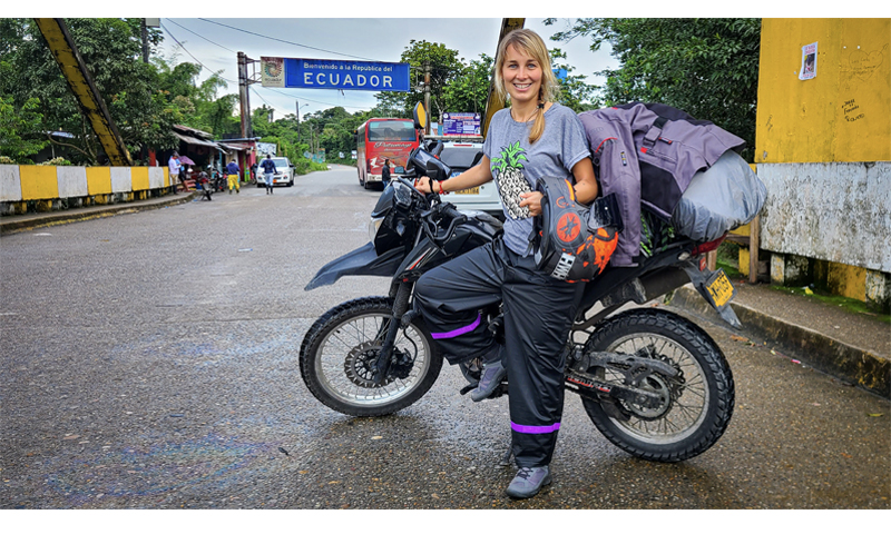 Uśmiechnięta kobieta opiera się o stojący za nią motocykl. W jednej ręce trzyma kask. Drugą opiera o kierownicę pojazdu. W tle ulica i niebieska tablica z napisem ECUADOR