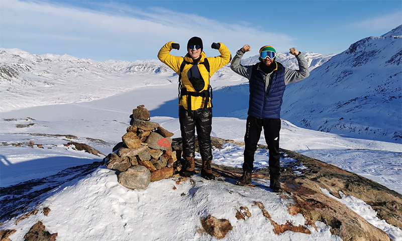 Dwóch mężczyzn w zimowych ubraniach stoi z uniesionymi ramionami na szczycie góry. W tle ośnieżone stoki gór